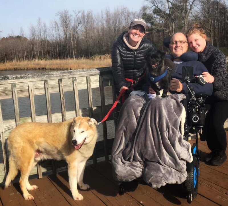 Two women, a man, and two dogs pose on a wooden outdoor walkway bordering a partially frozen body of water. All are dressed in winter clothes, and the trees in the background are bare, indicating winter. The man is in a wheelchair, his legs covered by a gray blanket. One dog sits on his lap, while the other stands, looking away from the camera. The women flank the man on each side.